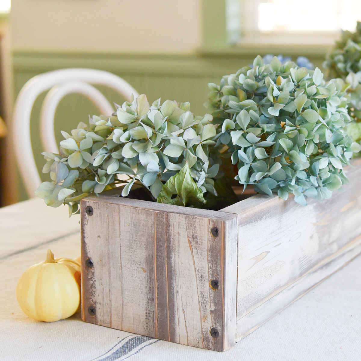 rustic diy box with dried blue hydrangeas in it and a small white pumpkin beside it, both setting on a grainsack tablecloth
