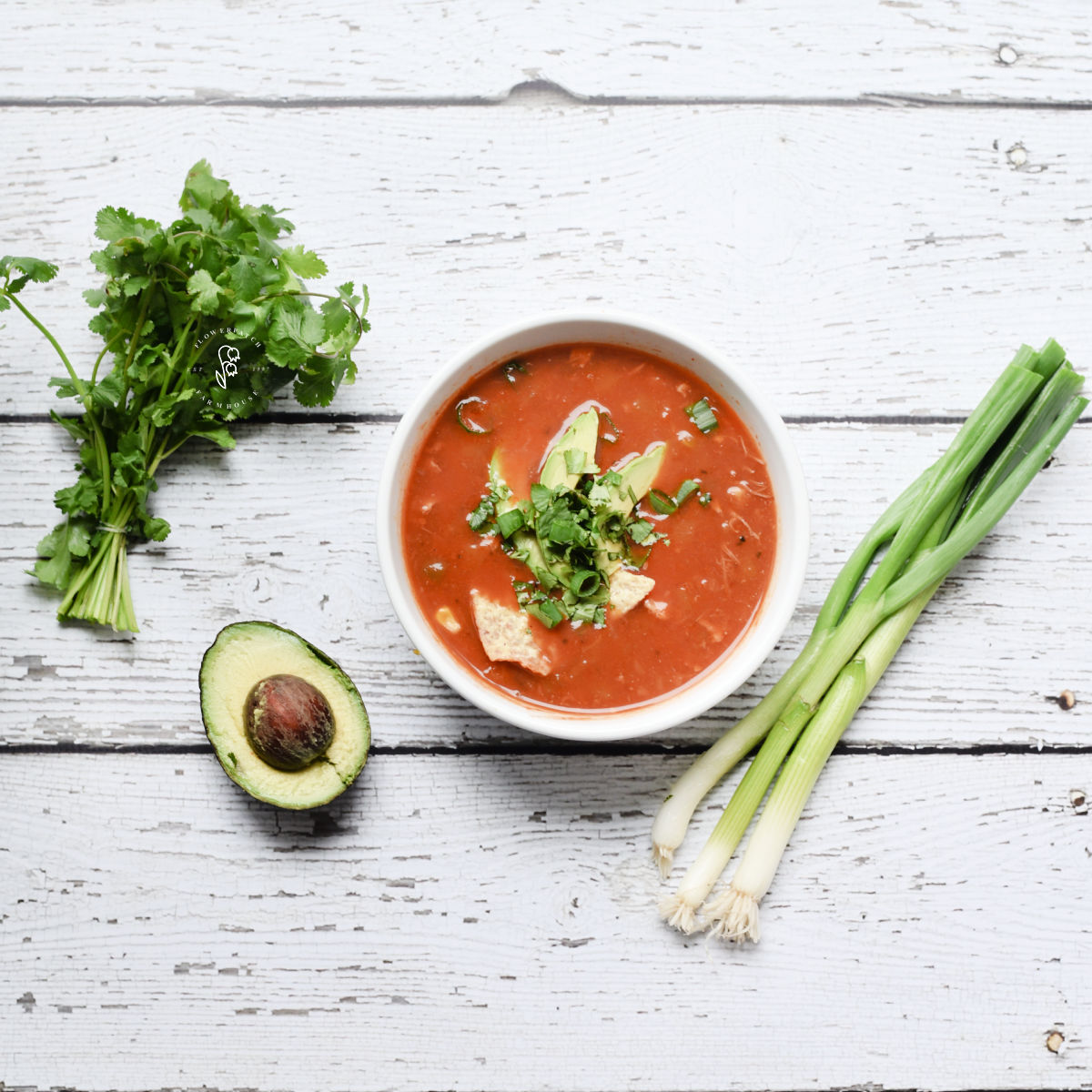 white bowl of chicken tortilla soup with green onions, half of an avocado and a bunch of cilantro flay layed on a white wood background