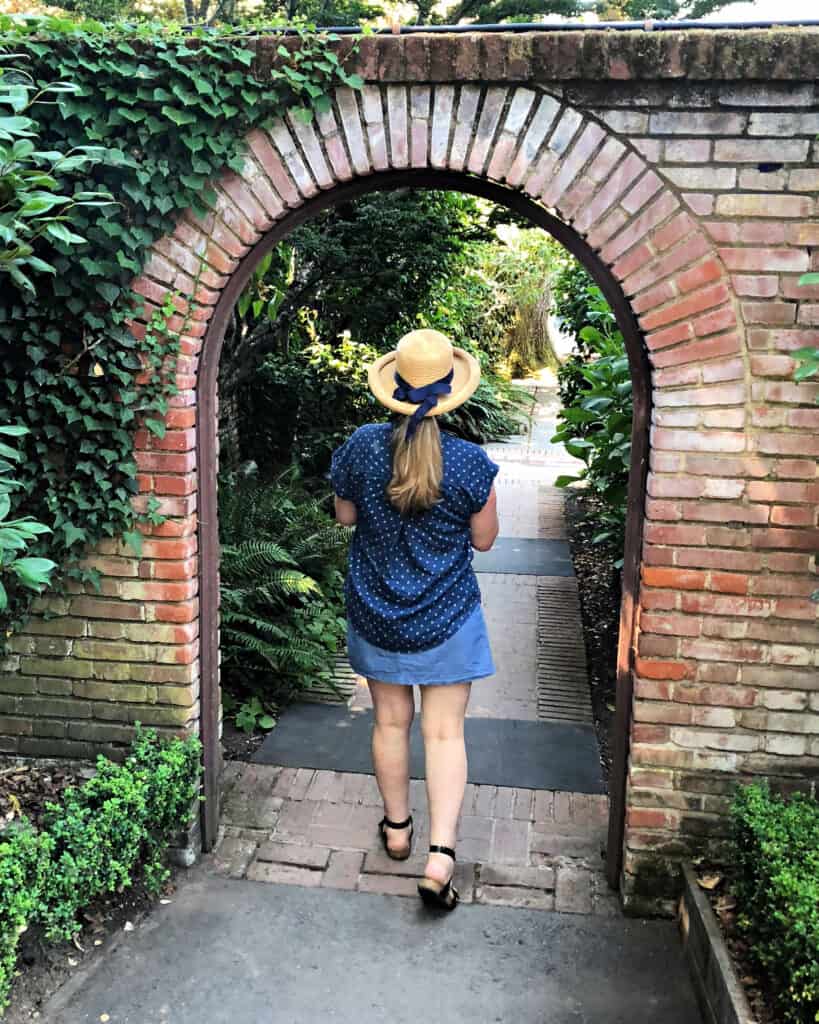 Pamela of Flower Patch Farmhouse dot com walking through an arched brick doorway into a lush garden