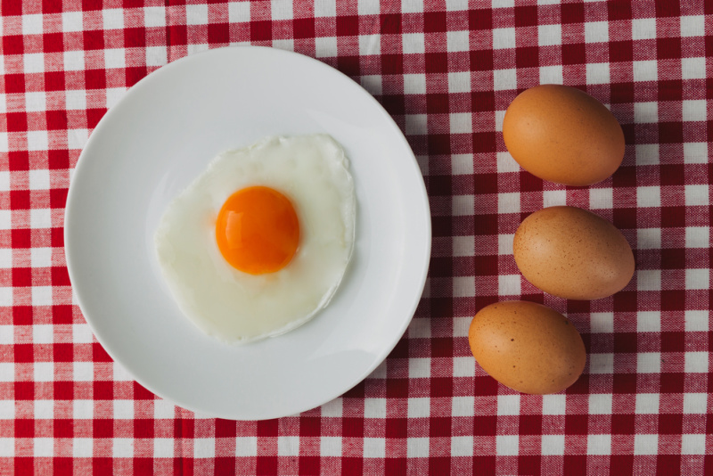 fried egg with orange yolk on a white plate, sitting on a red gingham tablecloth, three brown eggs sitting the the left