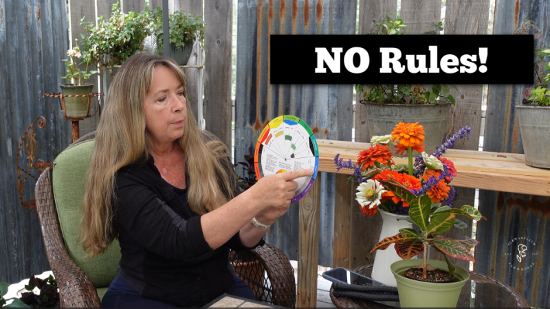 woman pointing to a color wheel, a vase of orange zinnias and white zinnias paired with blue salvias sit on a table beside her