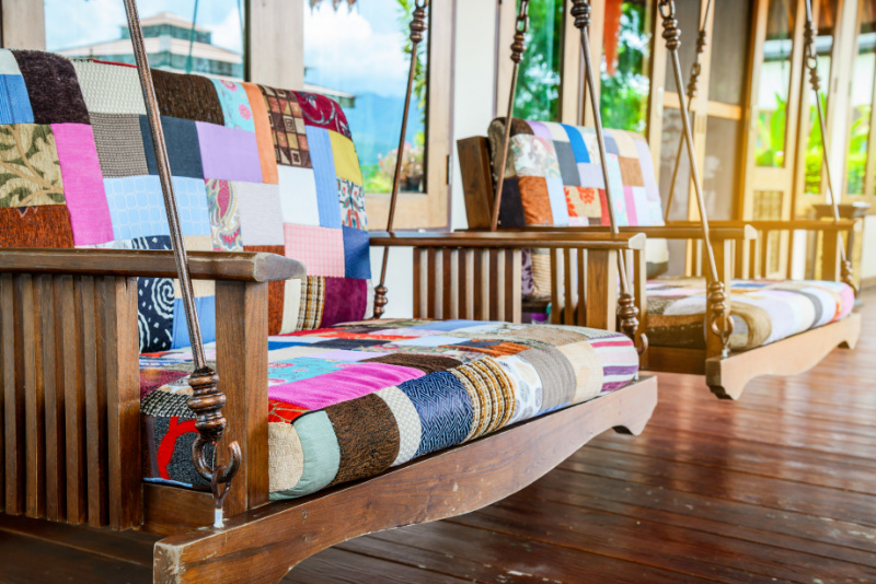 Two wooden porch swings with colorful patchwork cushions are suspended from the ceiling. The swings have a classic design with vertical slats and armrests. The vibrant fabric on the seat and backrest adds a cozy and eclectic touch to the porch area, which has wooden flooring and large windows in the background, offering a view of greenery outside.