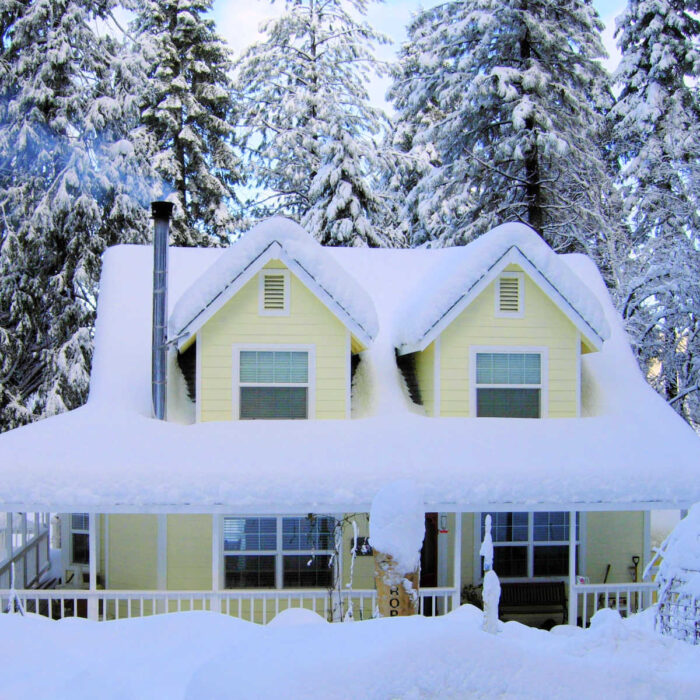 Yellow Cottage draped with snow with smoke coming from the chimney, evergreen trees in the background
